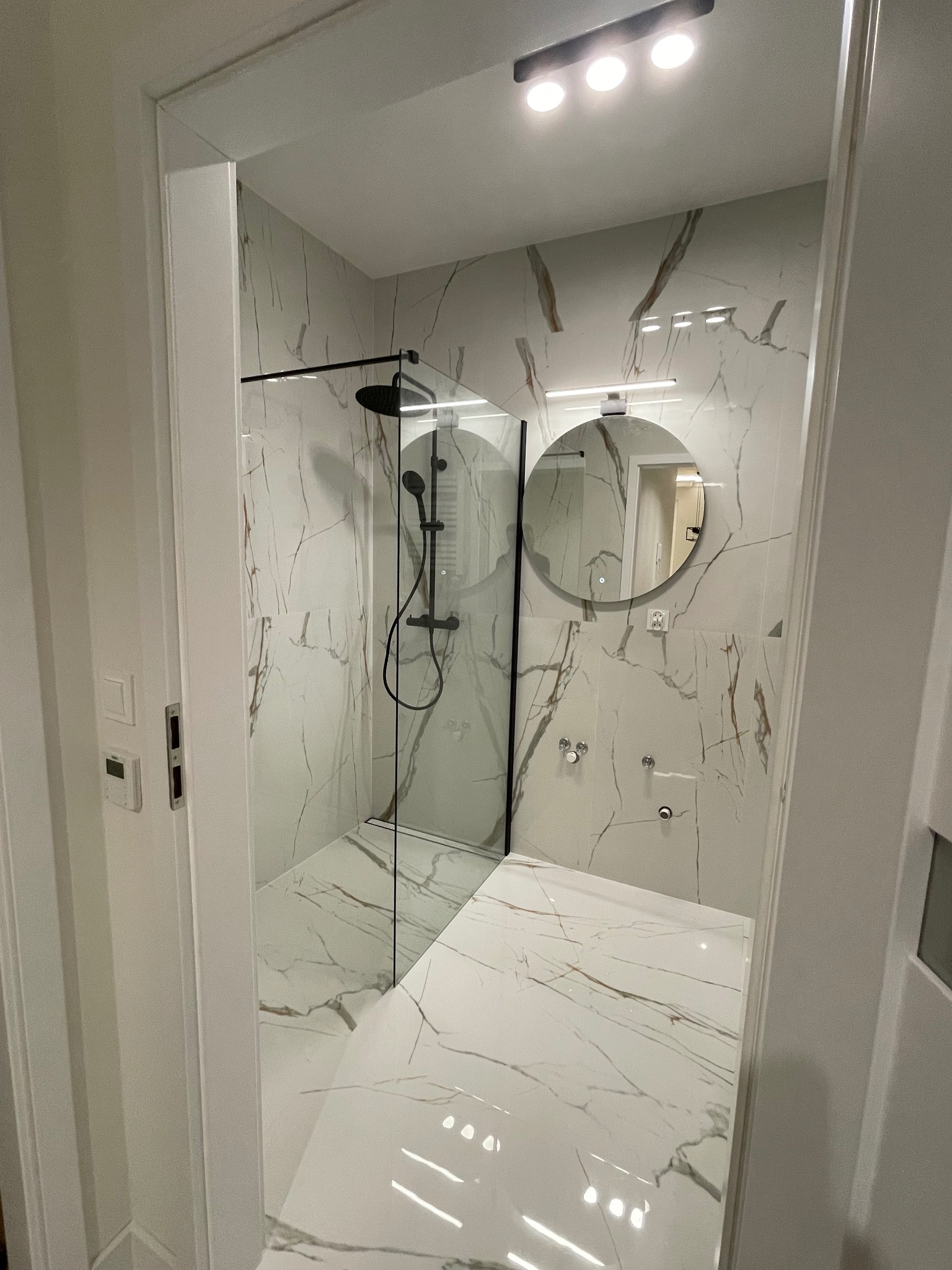 Nowoczesna łazienka z kabiną typu walk-in, wykończona białymi płytkami imitującymi marmur z brązowymi żyłami. Czarna armatura i okrągłe lustro dodają elegancji.