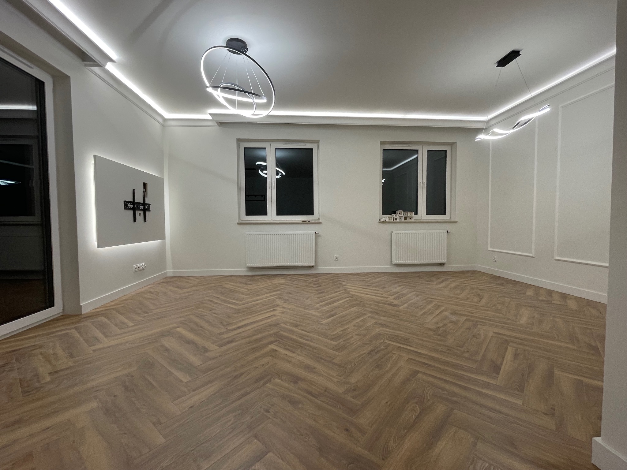 Nowoczesne wykończenie wnętrza: parkiet w jodełkę, białe ściany z dekoracyjnymi listwami, designerskie lampy i podświetlony uchwyt TV. Minimalizm i elegancja.