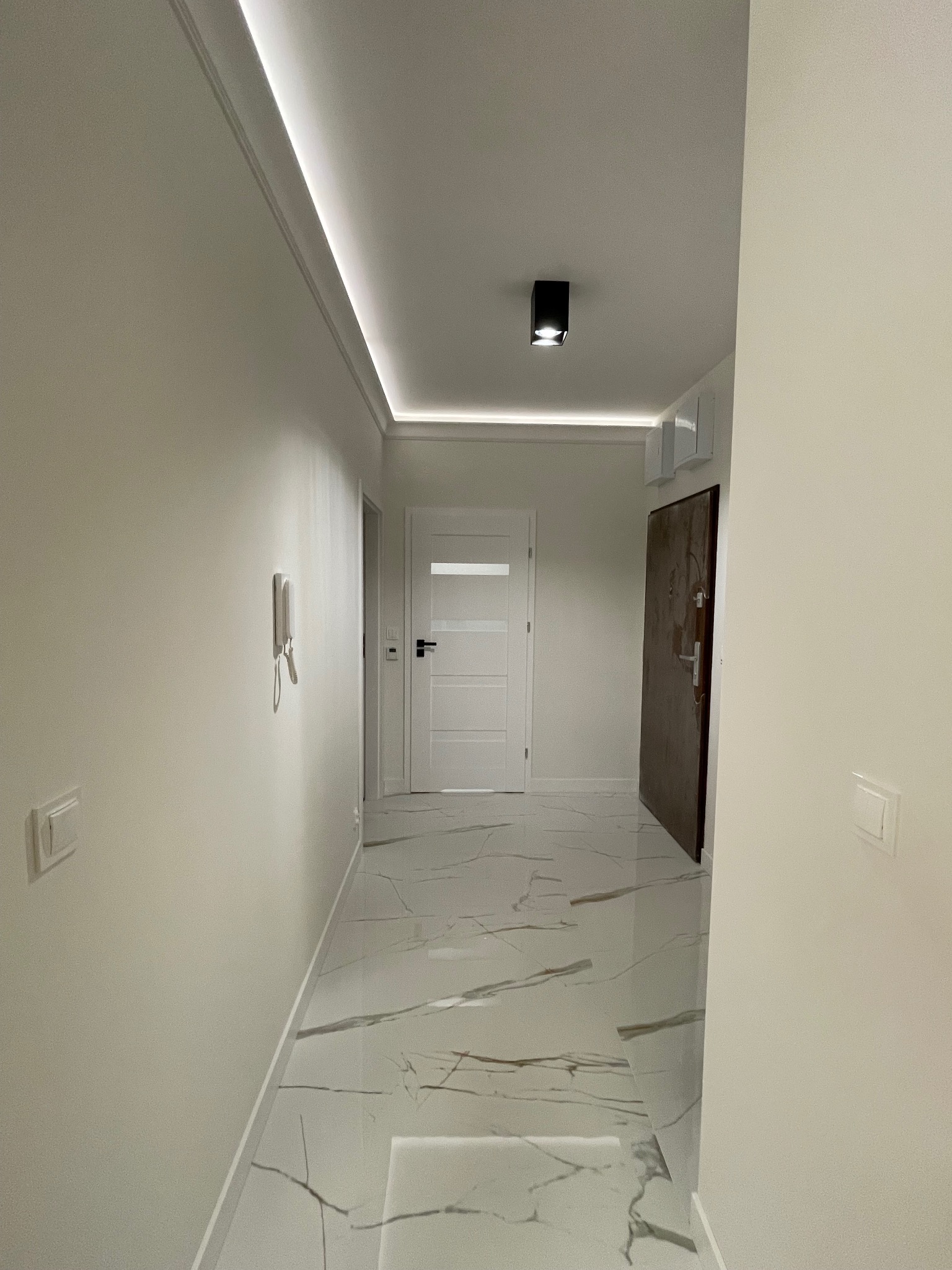 Jasne wnętrze korytarza z marmurową podłogą, białymi ścianami i nowoczesnym oświetleniem LED pod sufitem. Widoczne drzwi wejściowe i wewnętrzne.