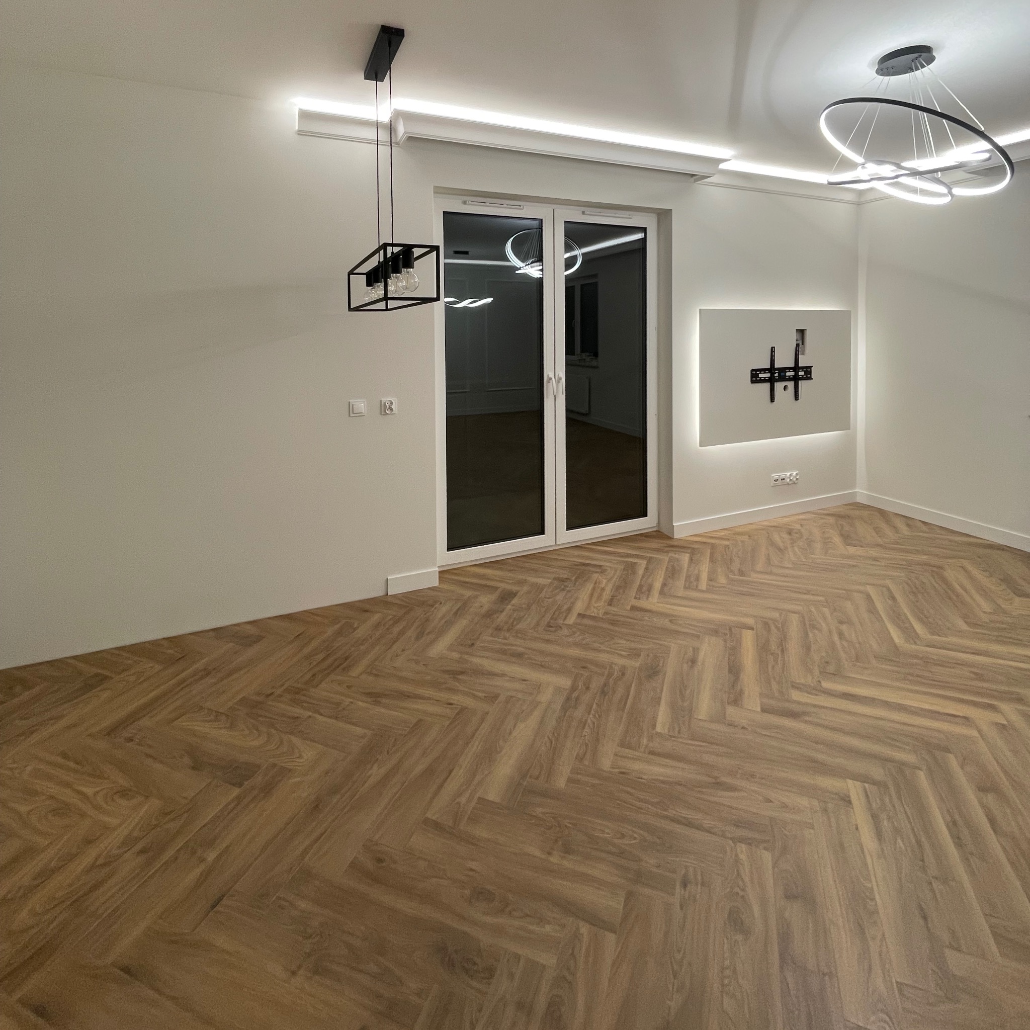 Nowoczesne wnętrze z parkietem w jodełkę, białymi ścianami i designerskim oświetleniem. Widoczne okno balkonowe i przygotowane miejsce na telewizor.