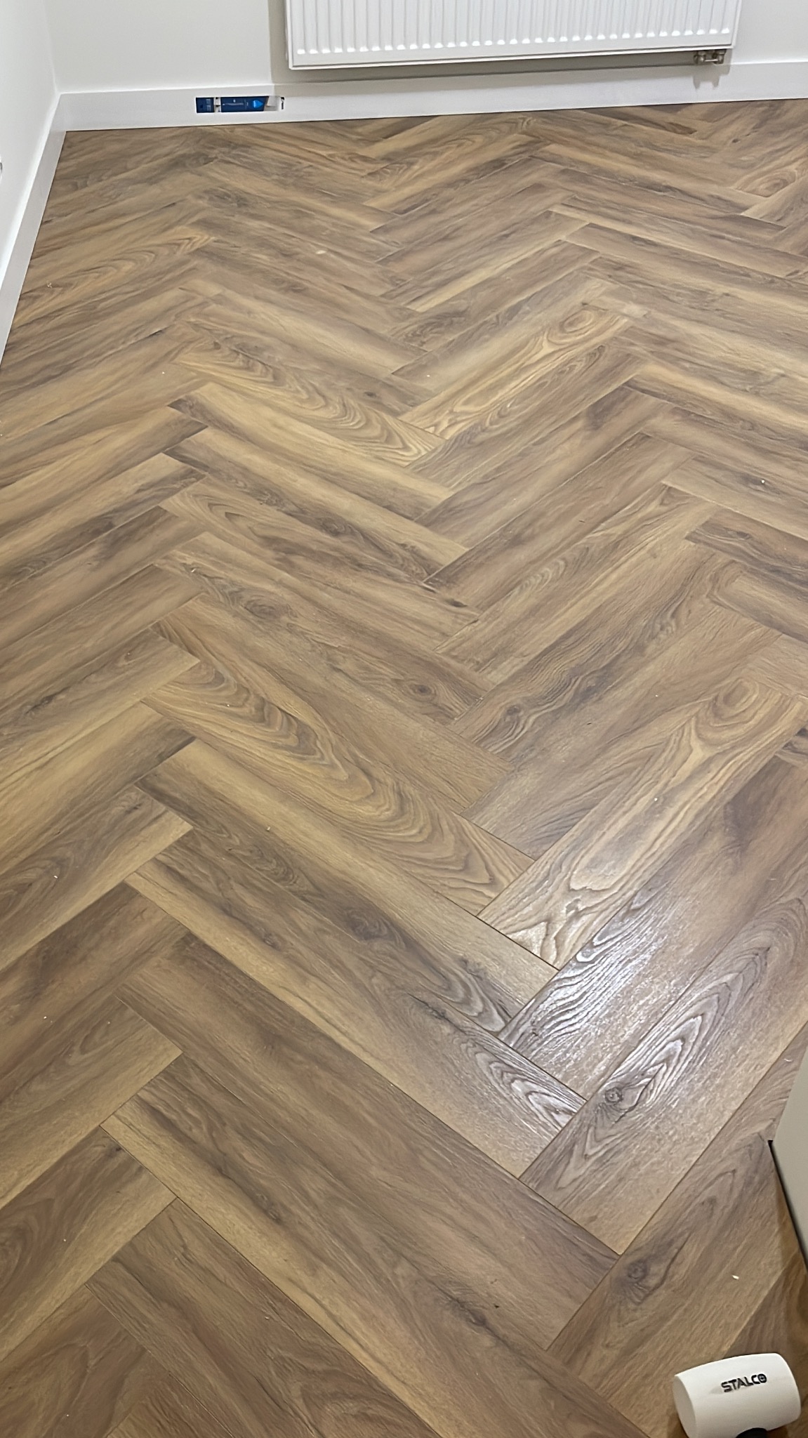 Ułożona podłoga z paneli w jodełkę, imitująca drewno, z widoczną listwą przypodłogową i grzejnikiem. Na podłodze leży biały odkurzacz.