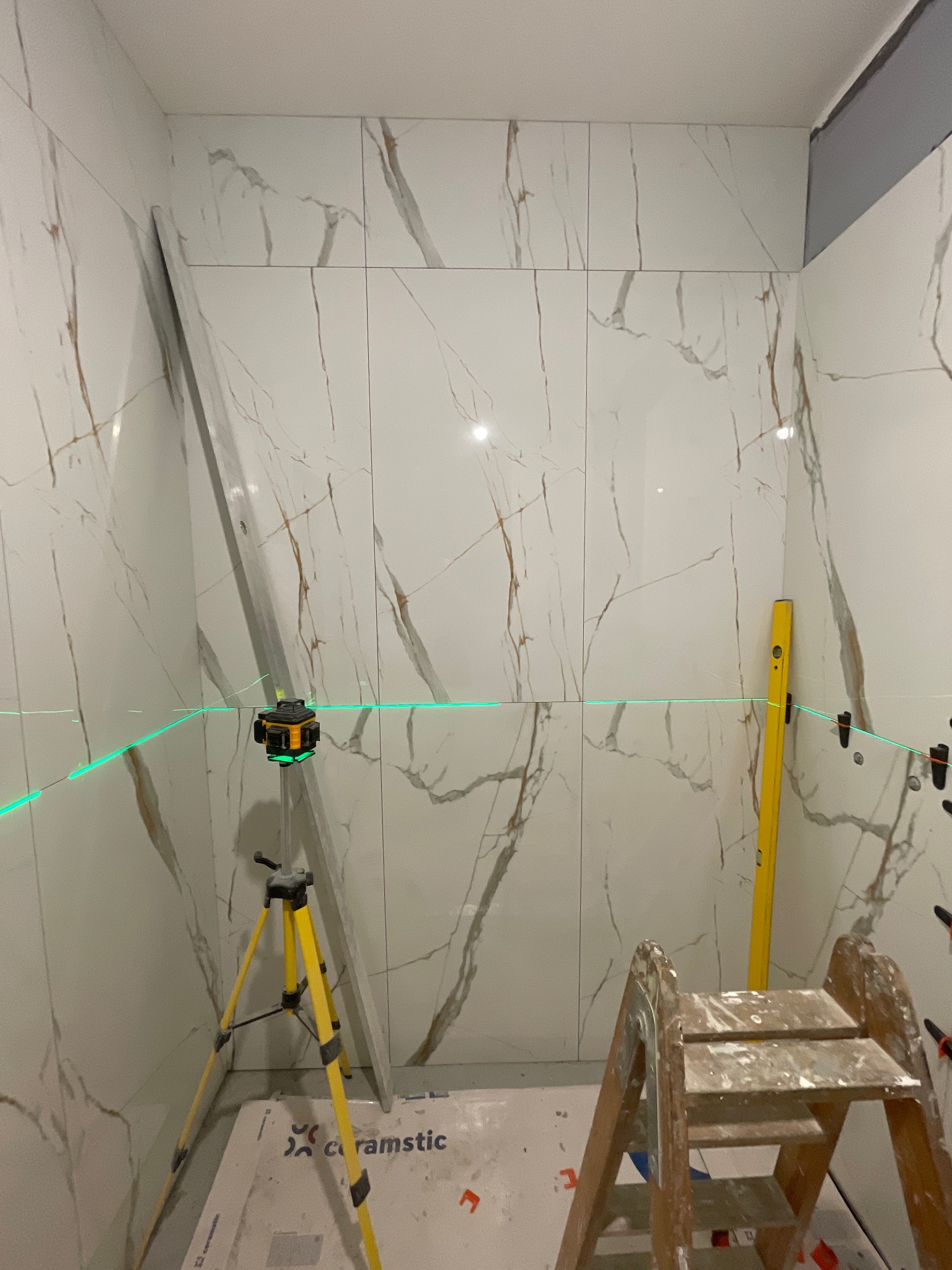 Układanie płytek imitujących marmur w łazience z użyciem poziomicy laserowej. Widoczna drabina malarska i poziomica. Etap prac wykończeniowych.