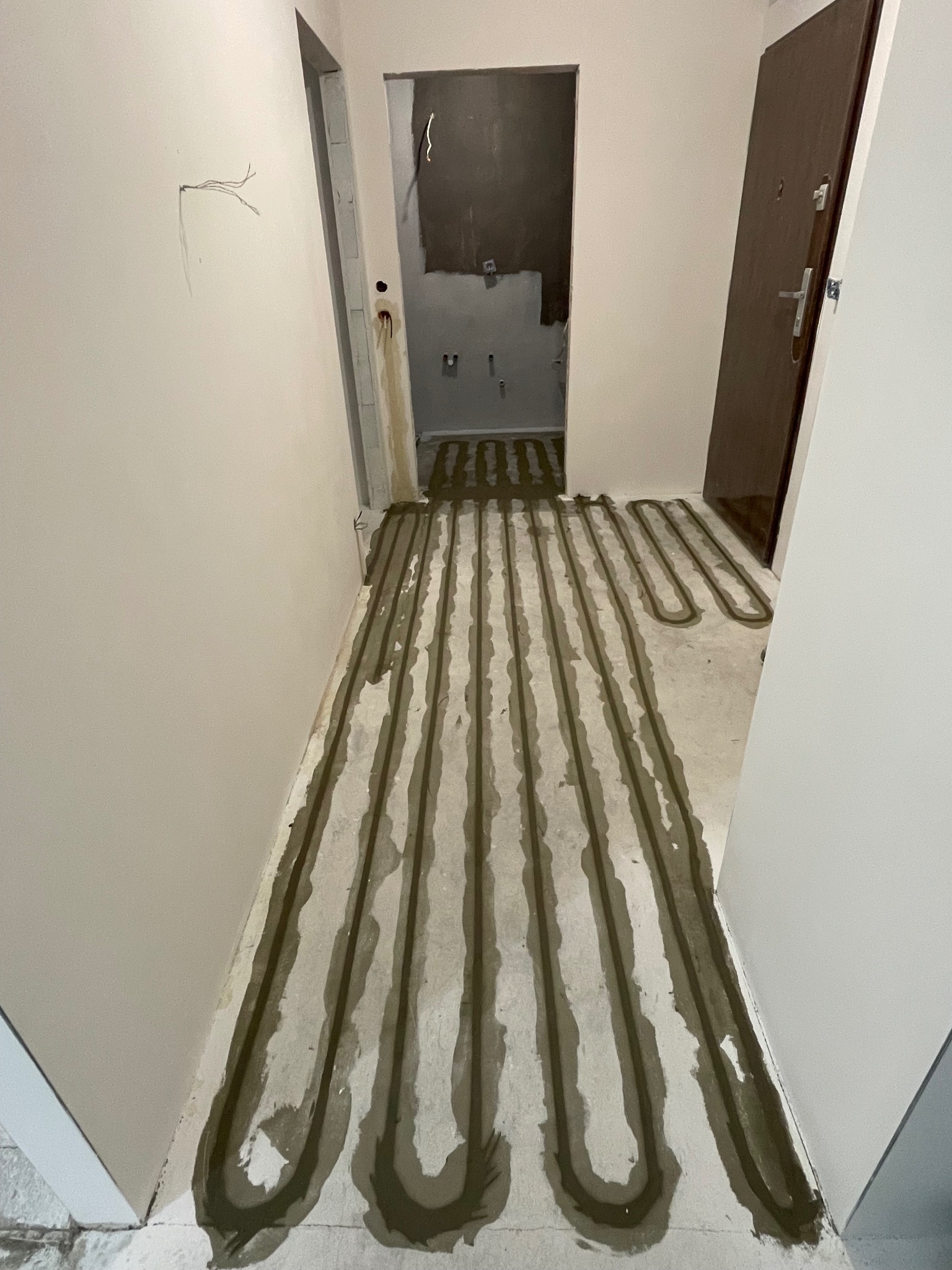 Wnętrze w trakcie remontu: ułożone rury ogrzewania podłogowego w jasnym korytarzu, widoczne przygotowania do wylewki. W tle wejście do łazienki i brązowe drzwi.