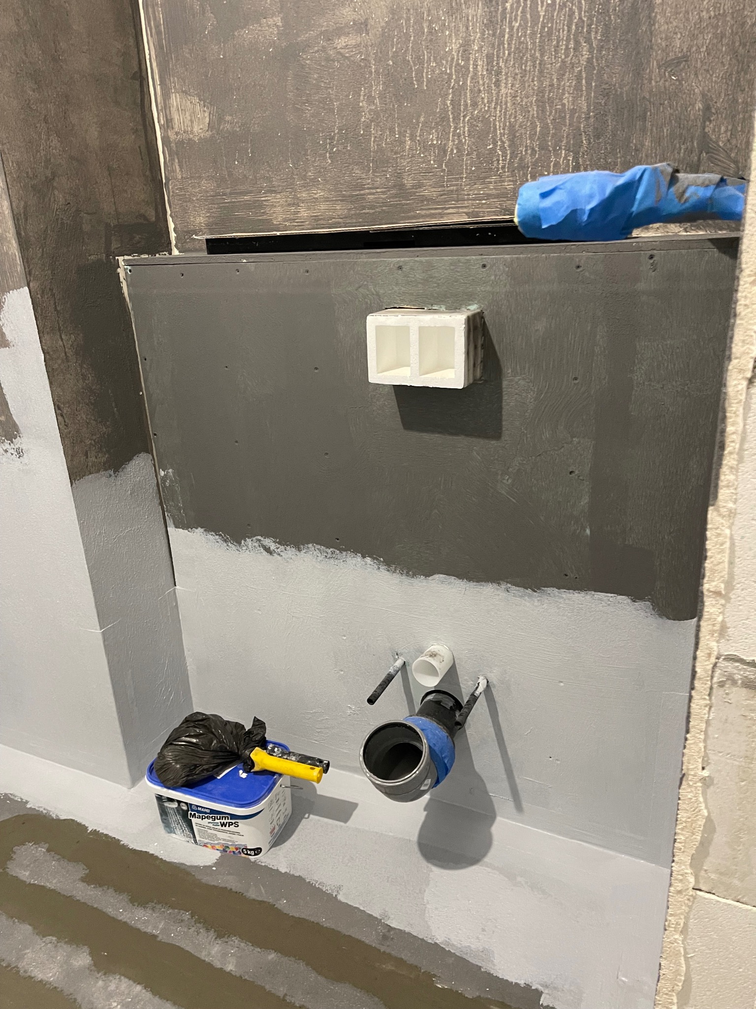 Przygotowanie łazienki do prac wykończeniowych: hydroizolacja ścian i podłogi, widoczne elementy instalacji i pojemnik z preparatem Mapegum WPS.
