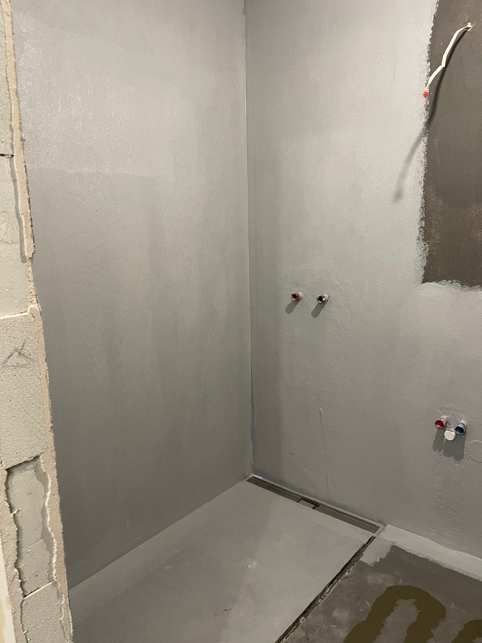 Szara, świeżo wykończona łazienka z odpływem liniowym. Widoczne przyłącza wodne na ścianie i fragment niezabezpieczonego przewodu elektrycznego.
