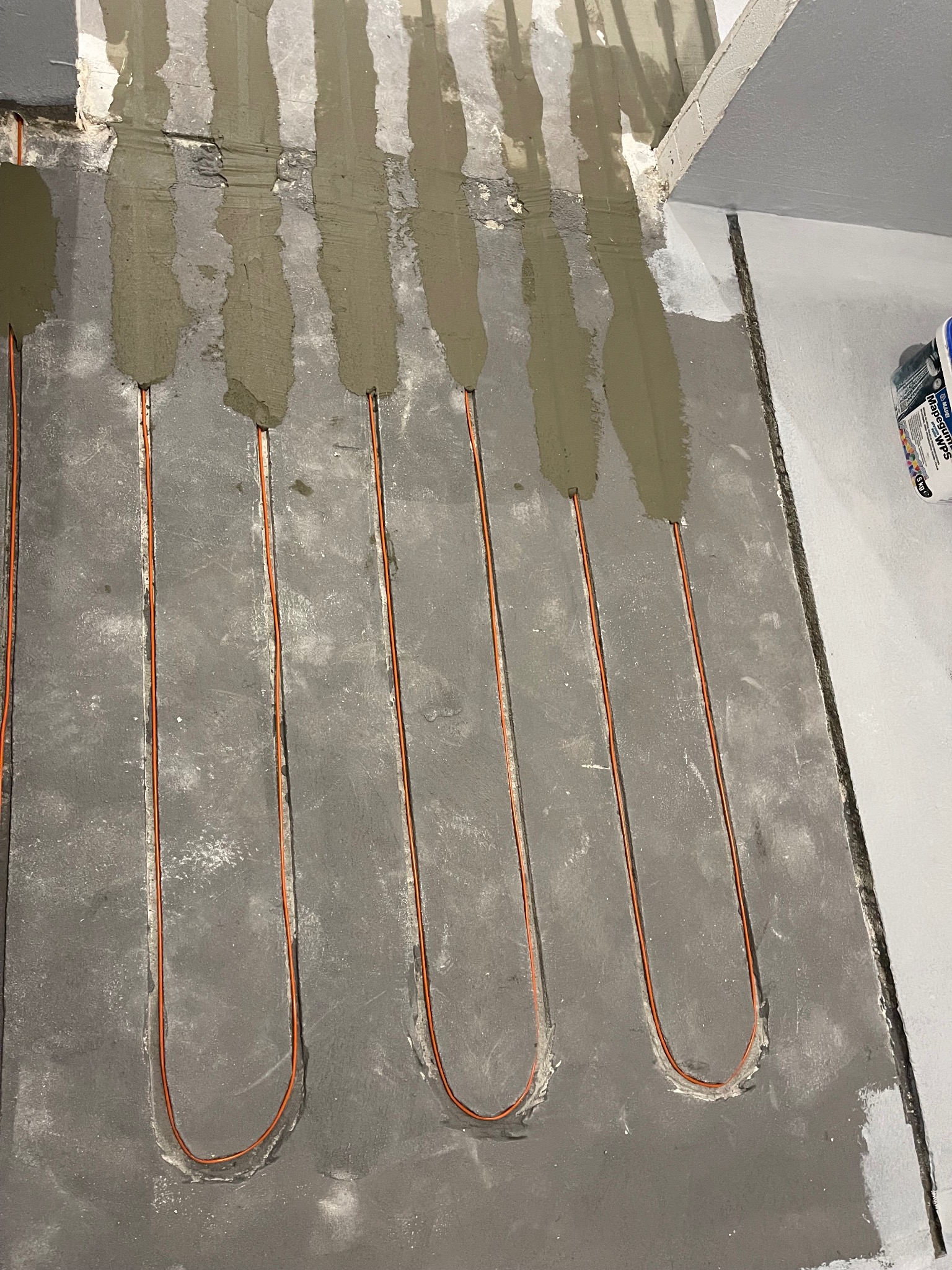 Montaż ogrzewania podłogowego: pomarańczowe kable grzewcze w szarym betonie, częściowo zalane zaprawą. Widoczne wiadro z preparatem gruntującym.