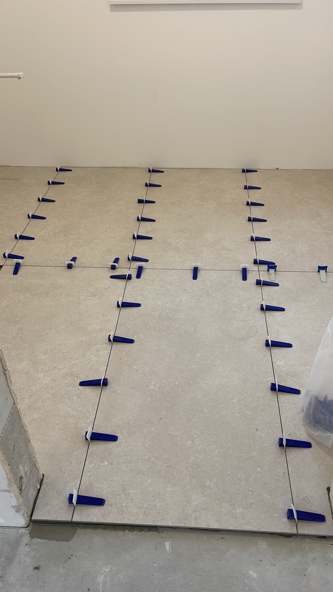 Ułożone płytki podłogowe z niebieskimi klipsami poziomującymi i czarnymi krzyżykami, widok z góry, jasne wnętrze, etap prac wykończeniowych.