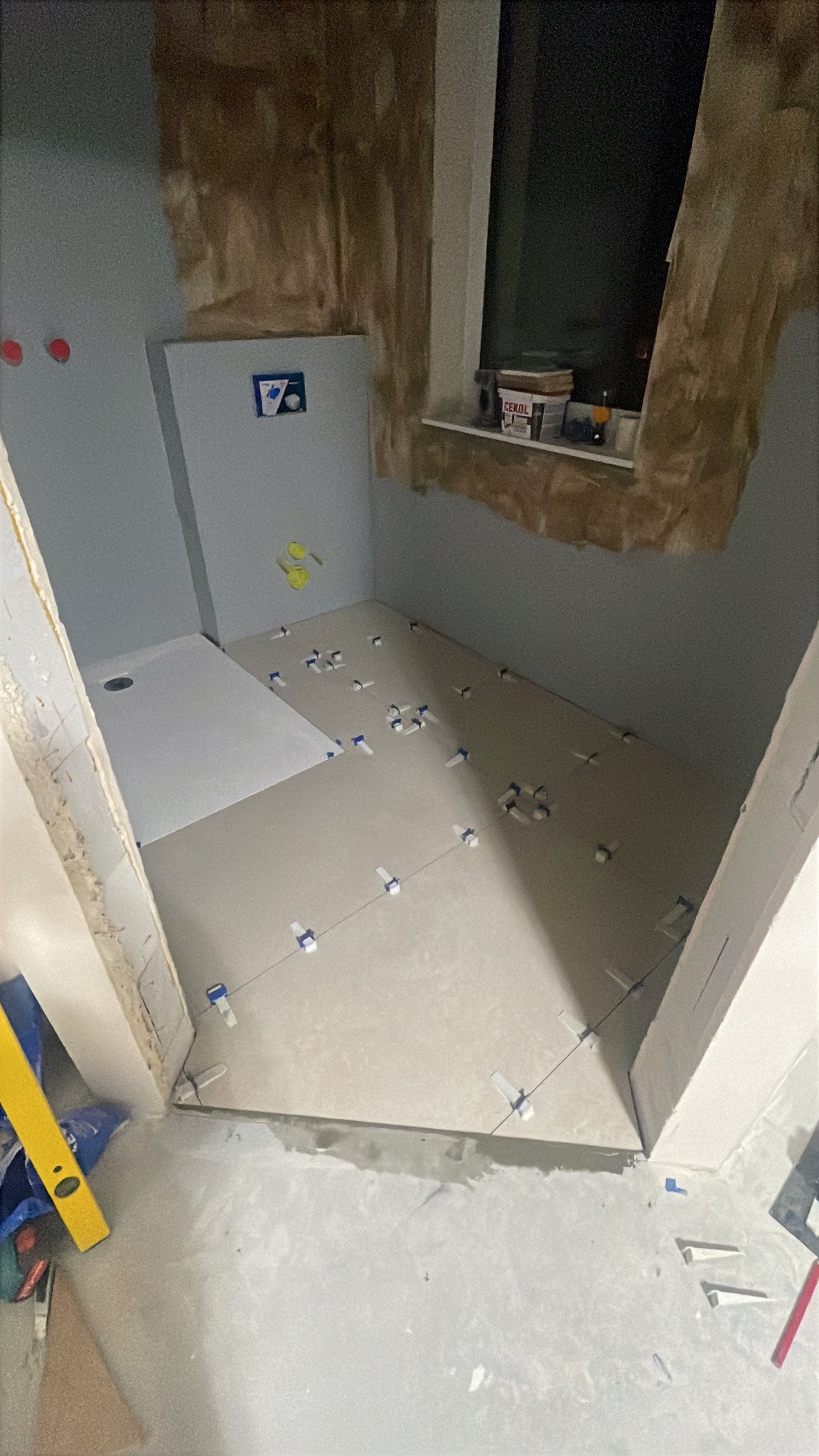 Łazienka w trakcie układania płytek podłogowych; widoczny brodzik, poziomica i krzyżyki glazurnicze, ściany przygotowane pod wykończenie.