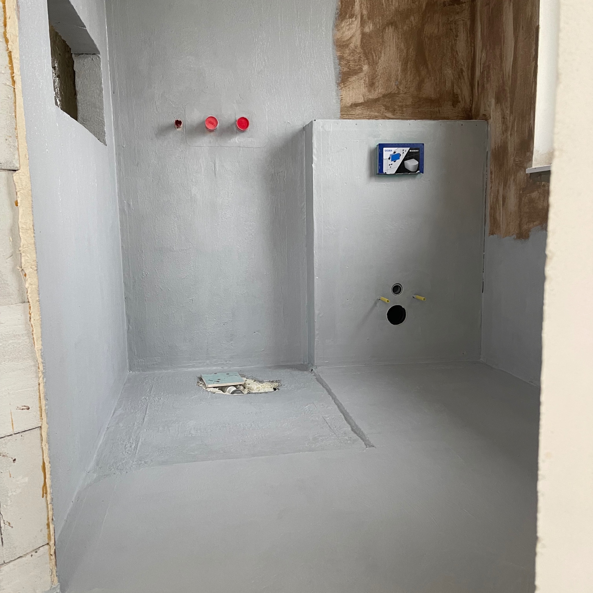 Szara hydroizolacja w łazience, widoczne rury, fragment ściany z instalacją podtynkową. Przygotowanie do dalszych prac wykończeniowych.