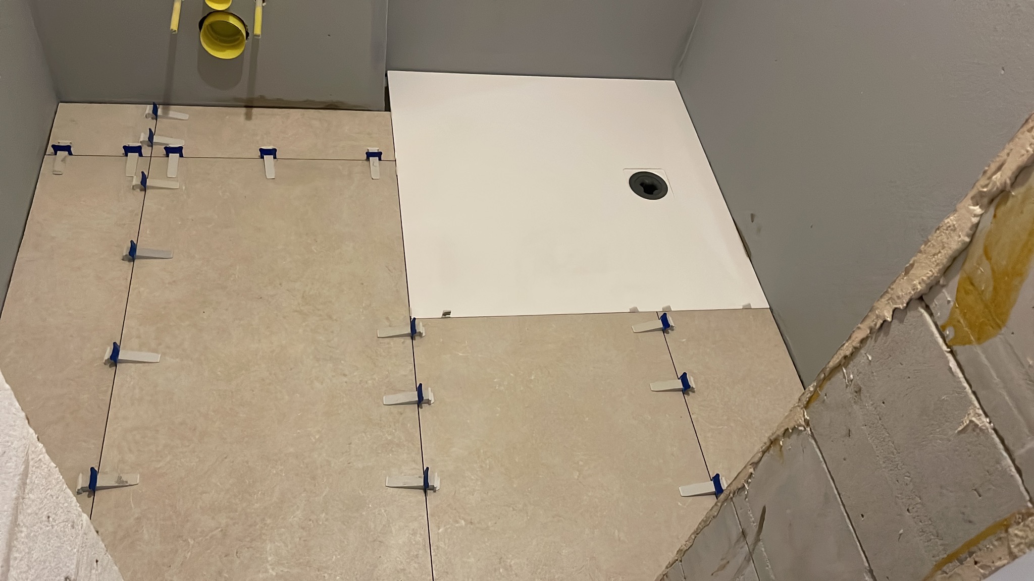 Układanie beżowych płytek w łazience z użyciem klipsów poziomujących. Częściowo ułożony biały brodzik z odpływem liniowym.