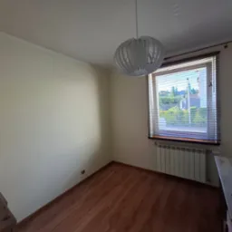 Jasne wnętrze pokoju z drewnianą podłogą i grzejnikiem pod oknem. Ściany są pomalowane na kremowo, a z sufitu zwisa nowoczesna, biała lampa.