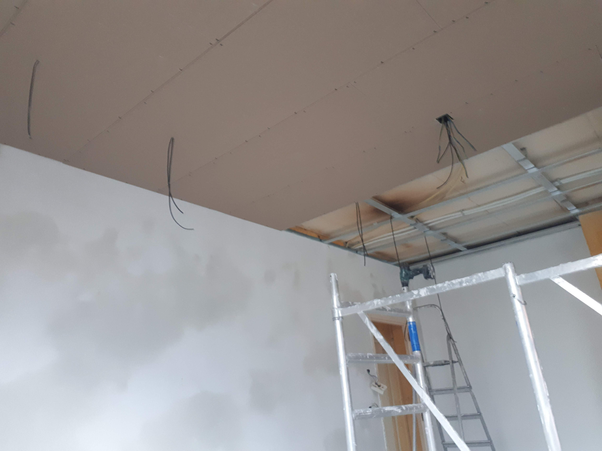 Montaż sufitu podwieszanego z płyty gipsowej, widoczne kable i rusztowanie. Częściowo wykończona ściana i płyty przymocowane do metalowego stelażu.