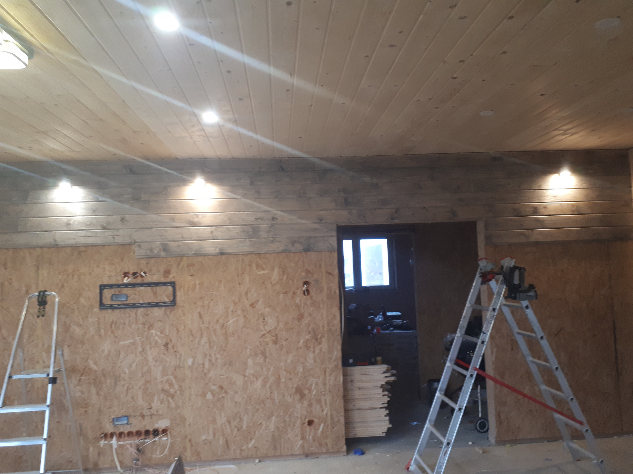 Wnętrze w trakcie wykańczania boazerią: drewniana podbitka sufitowa z zamontowanym oświetleniem punktowym, ściany z płyt OSB, drabiny i narzędzia. Widoczne przygotowania do dalszych prac.