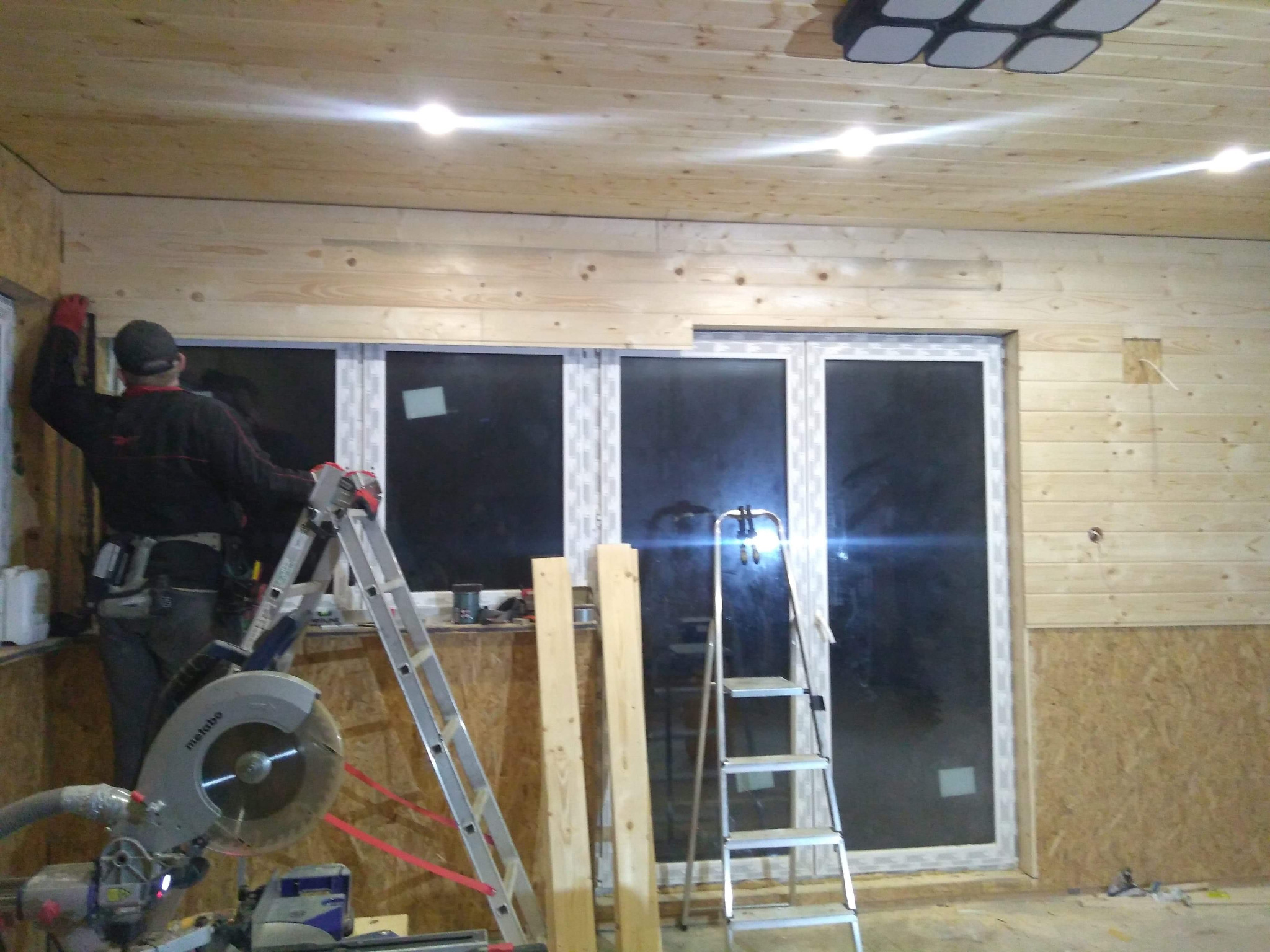 Prace wykończeniowe wnętrza z drewnianą boazerią: monter na drabinie dopasowuje deski. Widoczna piła, okno i oświetlenie sufitowe.