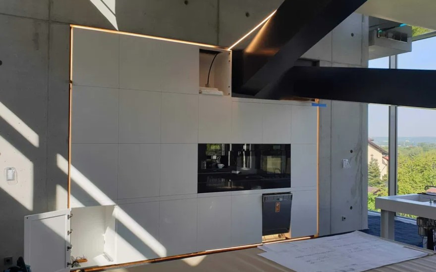 Biała zabudowa kuchenna z podświetleniem LED w nowoczesnym wnętrzu z betonowymi ścianami i dużym oknem z widokiem na zieleń. Na blacie leżą plany.