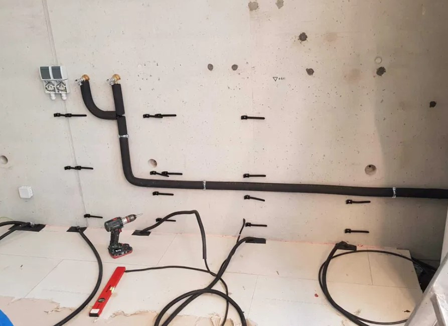 Montaż rur w izolacji termicznej na ścianie z betonu, widoczne uchwyty mocujące, poziomica i wiertarka. Etap prac instalacyjnych w surowym wnętrzu.