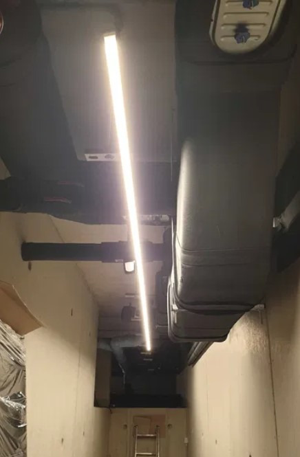 Długa, wąska lampa LED oświetla korytarz w piwnicy. Widoczne rury pod sufitem i drabina w oddali. Ściany w jasnym odcieniu beżu.