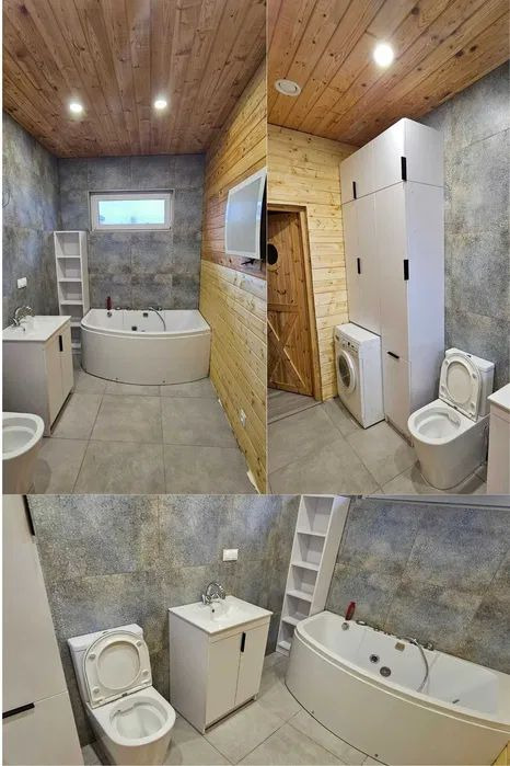 Wykończona łazienka z drewnianym sufitem i ścianą, szarą płytką, białą wanną z hydromasażem, toaletą, pralką i szafkami. Nowoczesny design z elementami rustykalnymi.