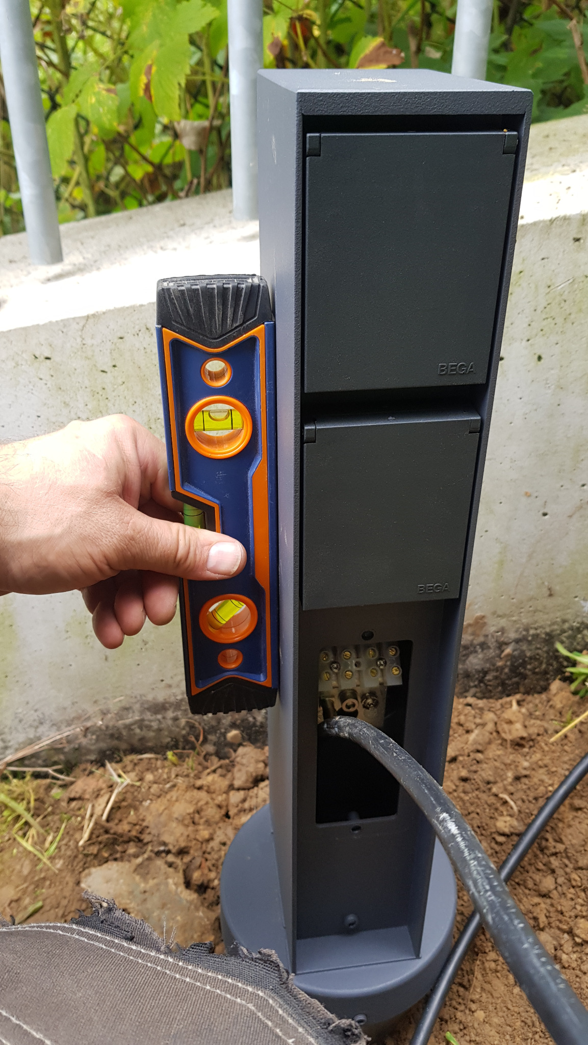 Instalacja zewnętrznej lampy Bega: elektryk poziomuje słup oświetleniowy, widoczne złącza elektryczne i kabel zasilający.