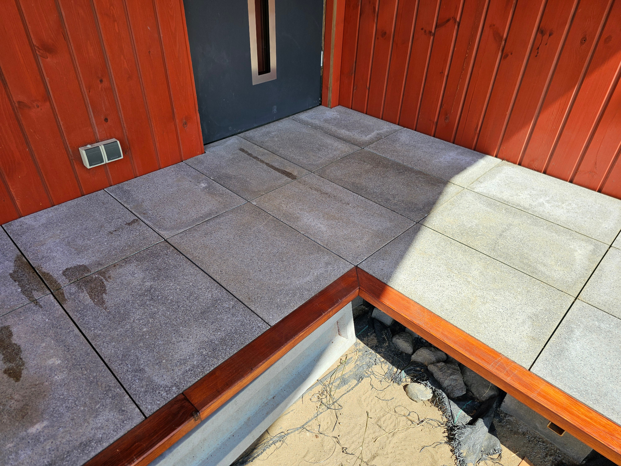 Nowa, szara kostka brukowa ułożona na tarasie z drewnianą obudową rabaty. Czerwone deski elewacyjne i szare drzwi wejściowe w tle. Widoczne ślady wody na kostce.