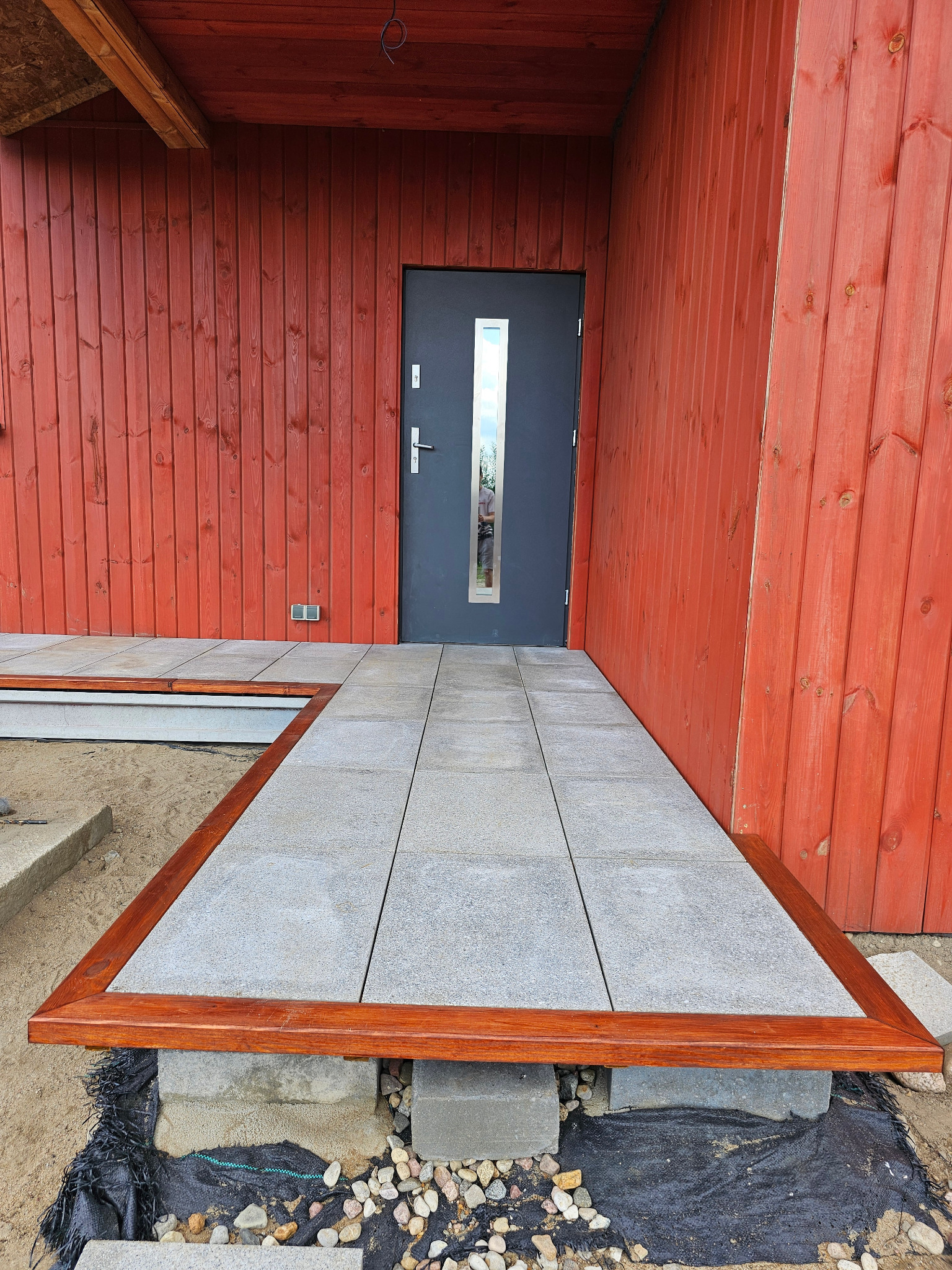 Nowe, szare płyty chodnikowe z drewnianą ramą prowadzące do drzwi wejściowych budynku z czerwoną elewacją. Widoczne bloczki betonowe pod płytami.