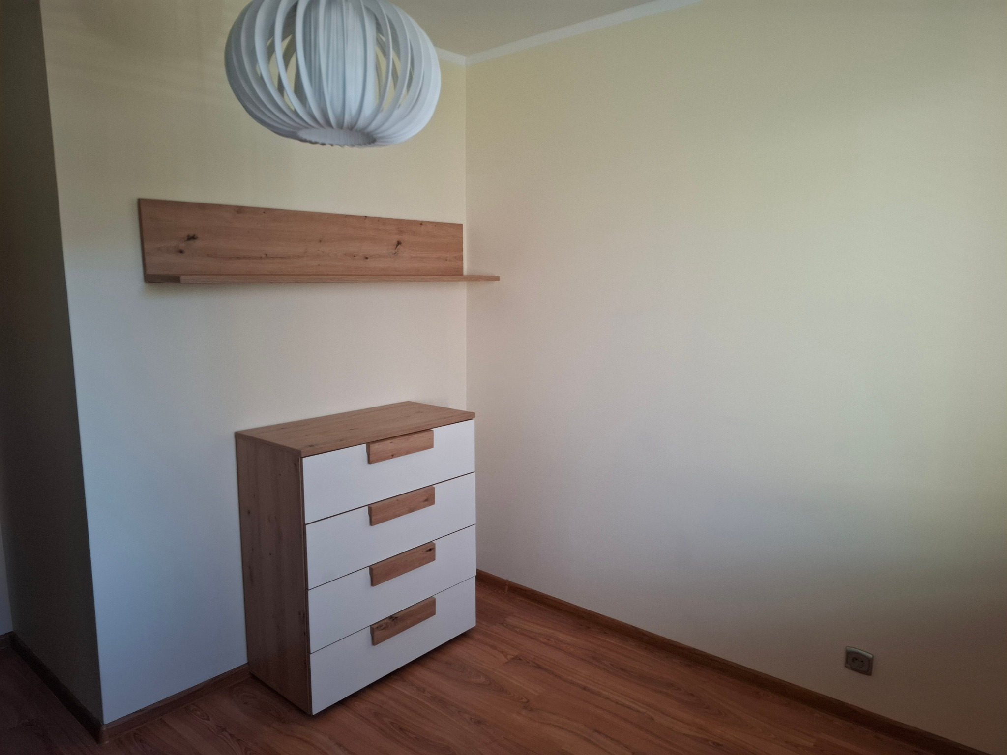 Komoda z czterema szufladami i półka z drewnianym blatem, wisząca lampa w pokoju z parkietem. Widoczny kontakt elektryczny na ścianie.