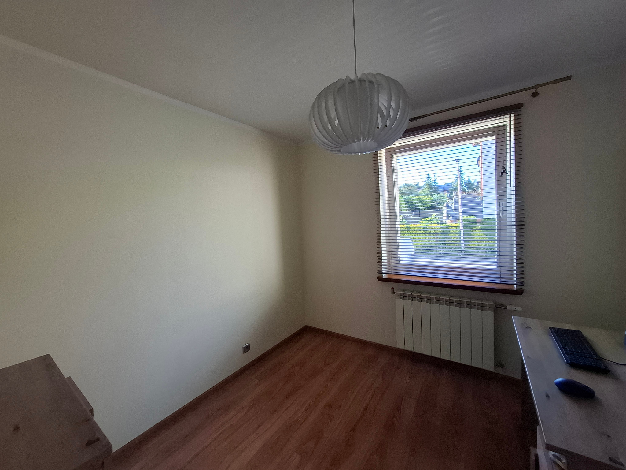 Jasne wnętrze pokoju z drewnianą podłogą i grzejnikiem pod oknem. Ściany są pomalowane na kremowo, a z sufitu zwisa nowoczesna, biała lampa.