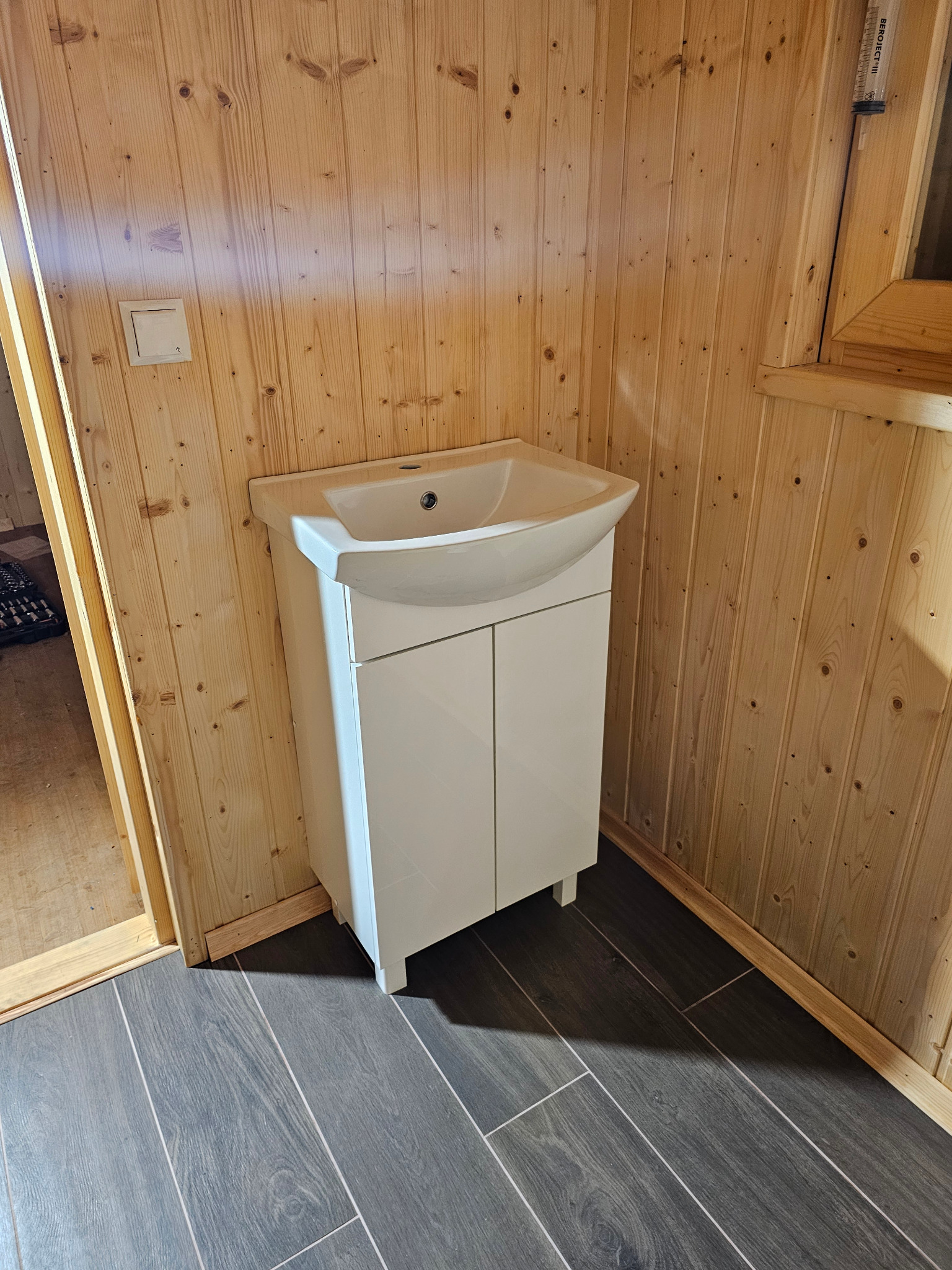 Biała szafka z umywalką w łazience wykończonej drewnem. Widoczne szare płytki podłogowe i fragment narzędzi w tle. Ujęcie z bliska, prezentujące detale wykończenia.