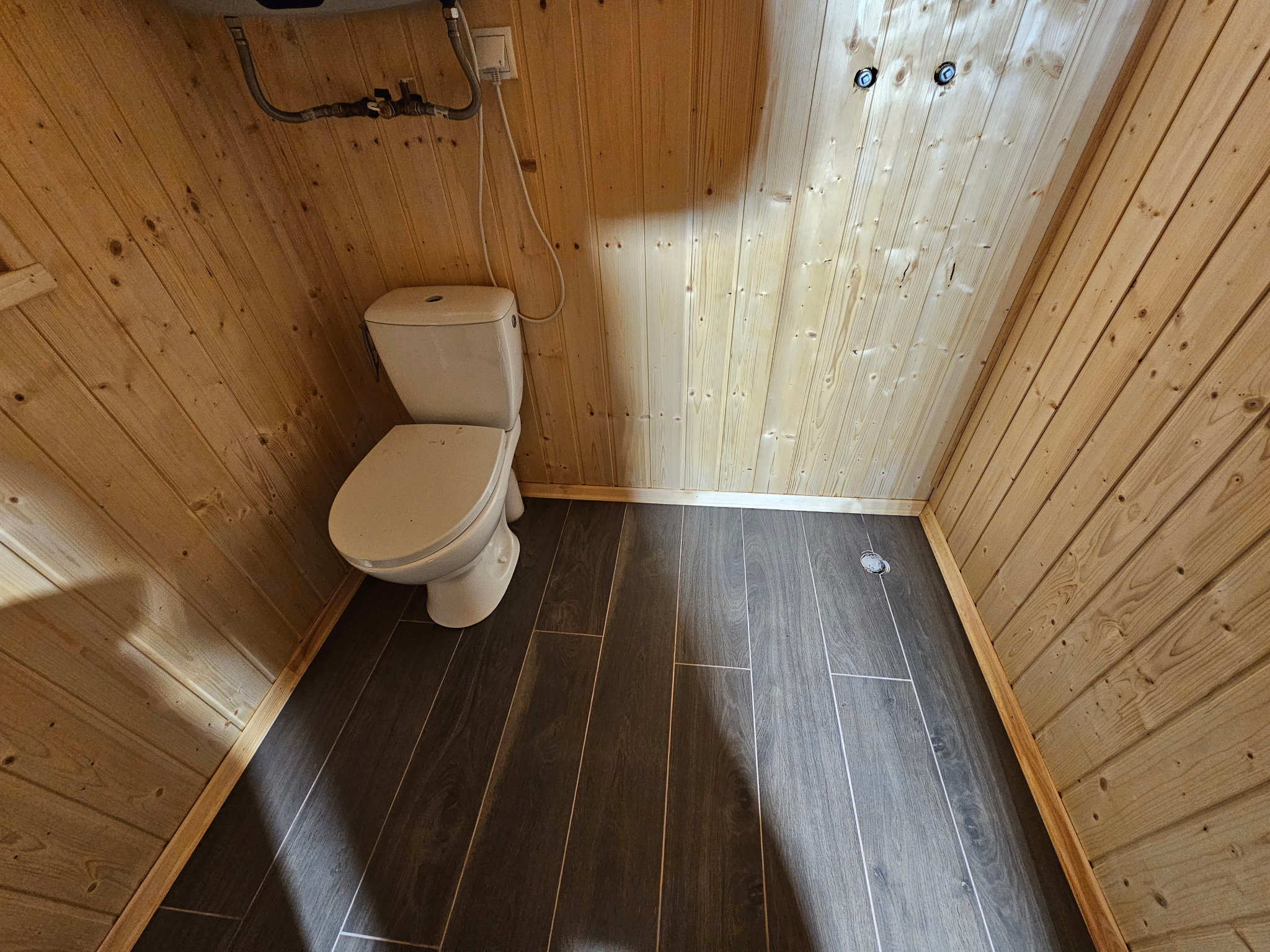 Drewnem wykończona toaleta z ciemną podłogą imitującą drewno. Widoczne przyłącza wodne i elektryczne. Ukończona aranżacja małej łazienki z WC.