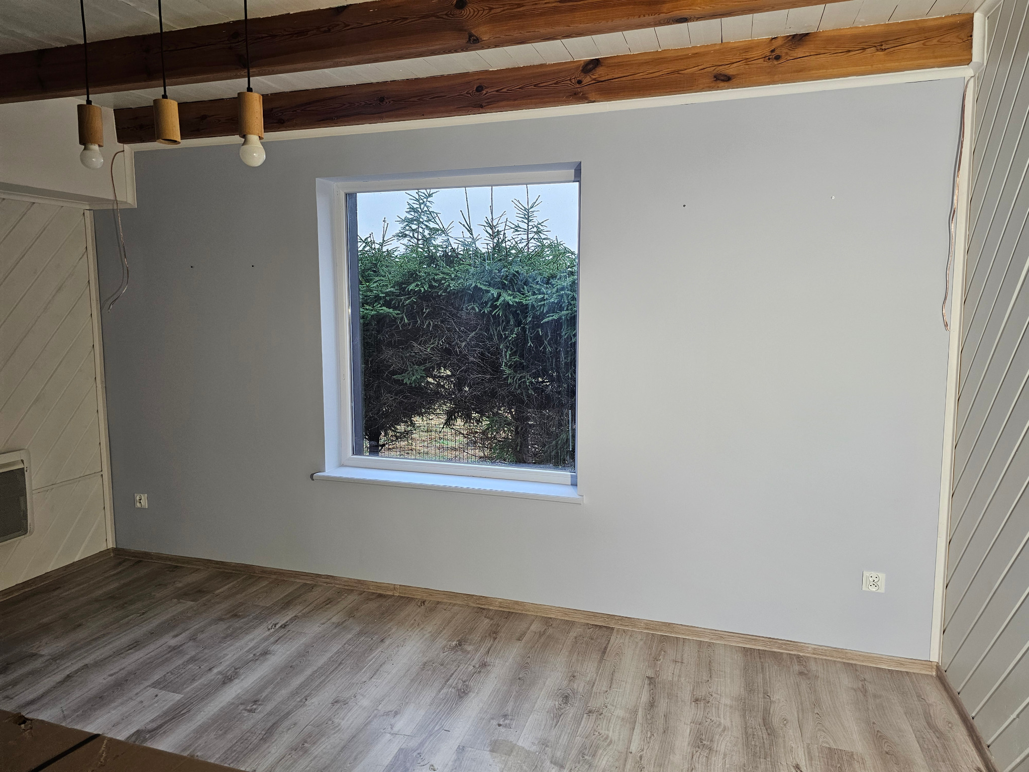Jasne wnętrze po malowaniu: szare ściany, drewniana podłoga, okno z widokiem na zieleń. Widoczne drewniane belki stropowe i nowoczesne lampy wiszące.