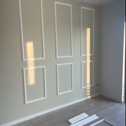 PAWEŁ-BUD Paweł Bulik - Ściana z zamontowanymi białymi panelami dekoracyjnymi w kształcie prostokątów, na podłodze leżą listwy przypodłogowe i elementy paneli, oświetlone promieniami słońca.