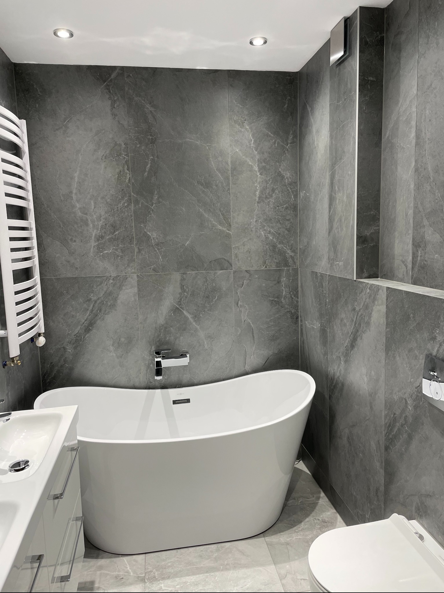 Nowoczesna łazienka w szarościach: wolnostojąca wanna, grzejnik drabinkowy i umywalka z szafką. Płytki imitujące marmur na ścianach i podłodze.