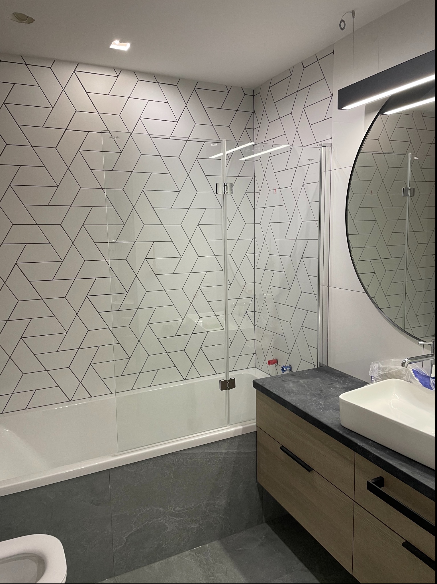 Nowoczesna łazienka po remoncie: białe płytki z geometrycznym wzorem, szklany parawan nawannowy, okrągłe lustro i drewniana szafka pod umywalką.