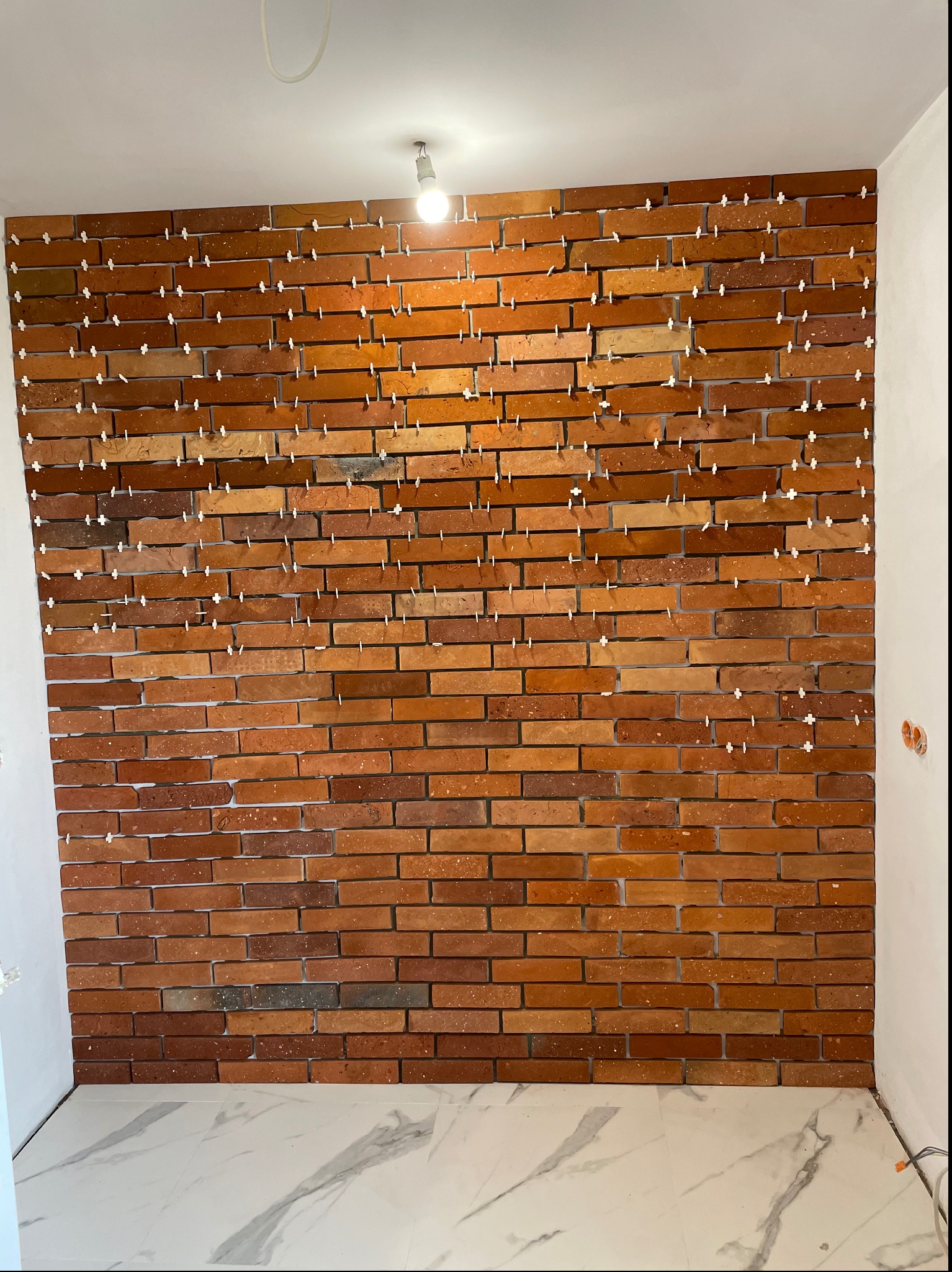 Ściana z cegieł klinkierowych w trakcie montażu, z widocznymi krzyżykami dystansowymi. Biała marmurowa podłoga, surowy stan wykończenia.