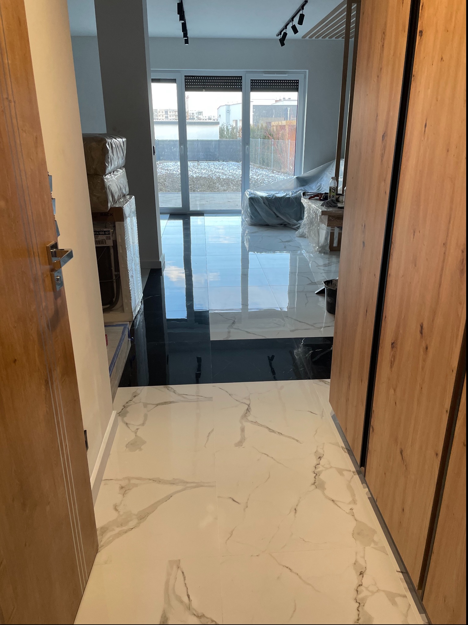 Wykończony fragment podłogi z białych i czarnych płytek w nowoczesnym wnętrzu. Widoczne drzwi z drewnianymi okładzinami i okno balkonowe z widokiem na zewnątrz.