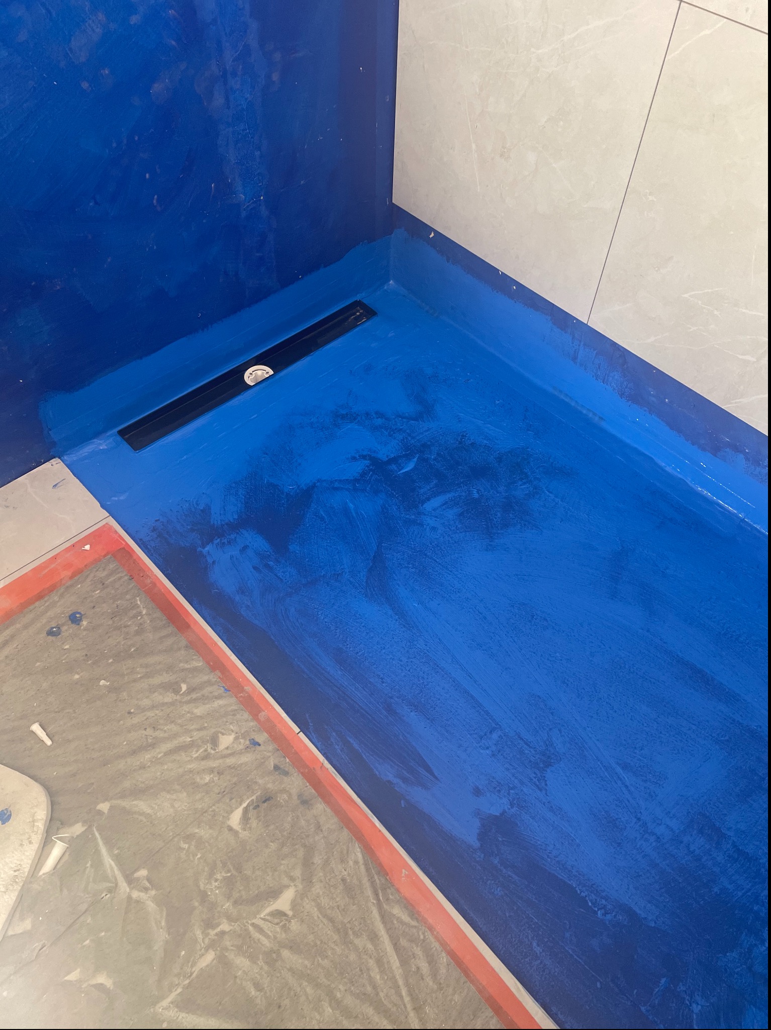 Hydroizolacja w kolorze kobaltowym, z odpływem liniowym w kabinie prysznicowej. Prace w trakcie realizacji w Środzie Wielkopolskiej. Widoczne płytki na ścianie.