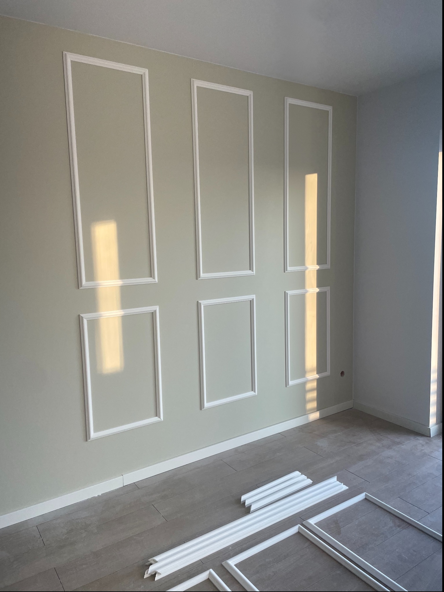 Ściana z zamontowanymi białymi panelami dekoracyjnymi w kształcie prostokątów, na podłodze leżą listwy przypodłogowe i elementy paneli, oświetlone promieniami słońca.