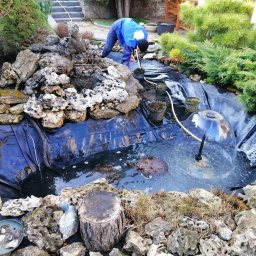 AQUAPRUD - Prace wykończeniowe wokół oczka wodnego: układanie kamieni, sadzenie roślin. W tle schody i roślinność ogrodowa.