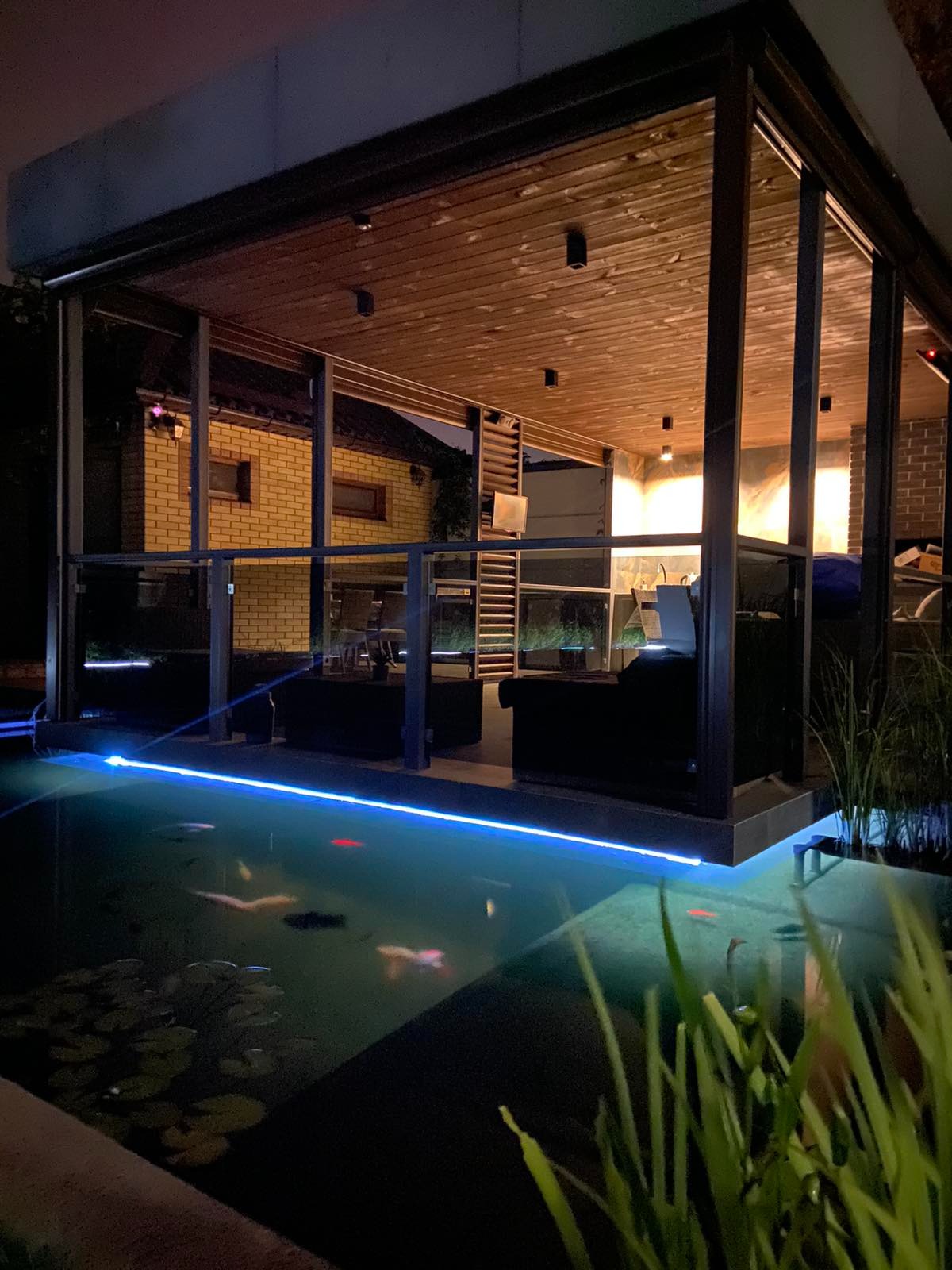 Oświetlone oczko wodne z rybami koi, liliami wodnymi i nowoczesną altaną z drewnianym sufitem, widoczne zarośla w tle, nocne ujęcie.