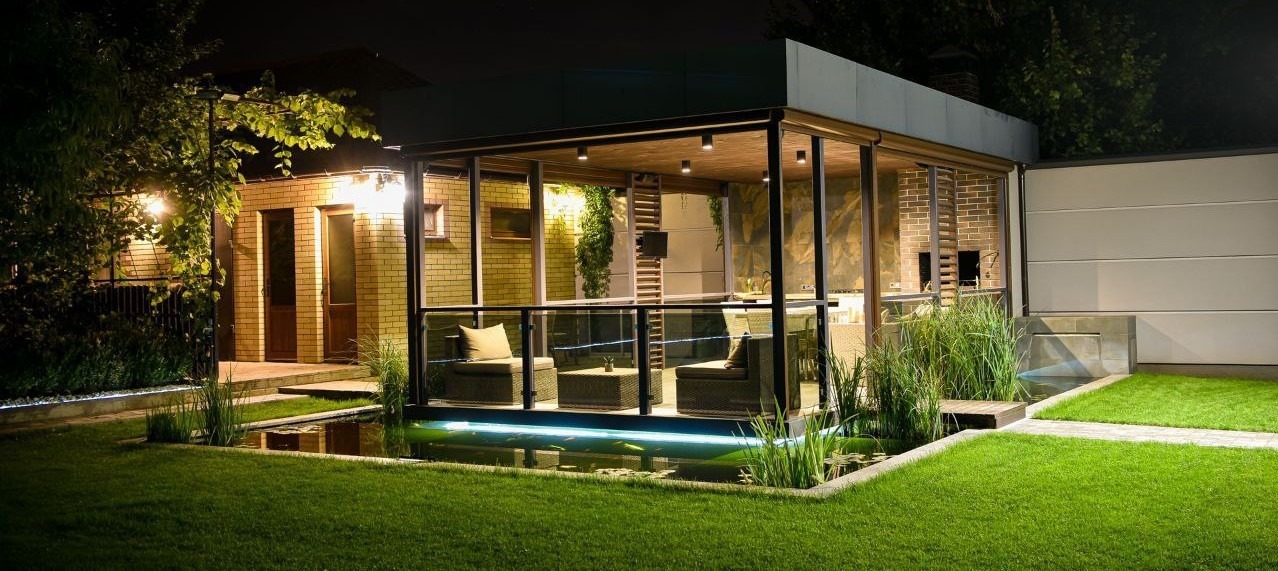 Oświetlone oczko wodne z roślinnością i nowoczesną altaną wypoczynkową na posesji. W tle ceglany budynek i jasny mur. Całość w nocnej scenerii.