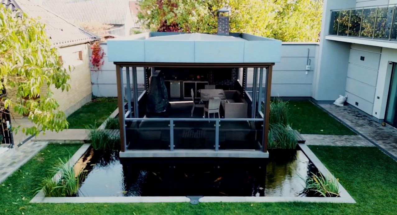 Nowoczesne oczko wodne z altaną na prywatnej posesji, widok z góry. Woda otoczona zielonym trawnikiem, w tle budynki mieszkalne i drzewa.