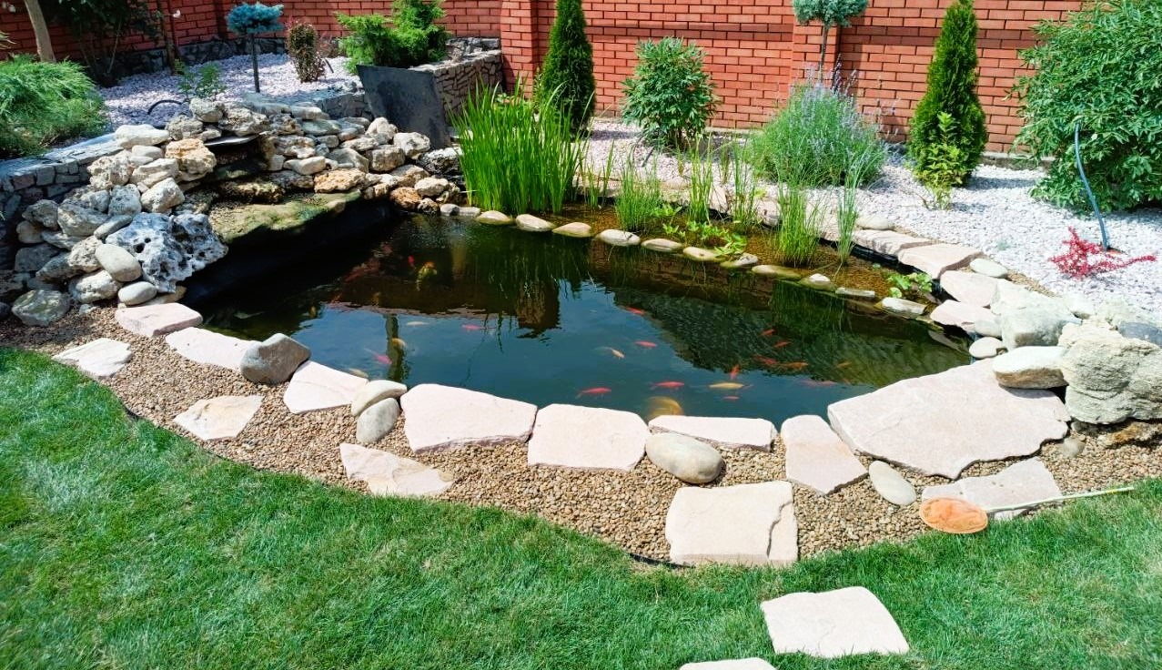 Ekostaw z wodospadem, obrzeża z kamieni i żwiru, naturalna filtracja, roślinność wodna, karasie ozdobne i karpie Koi, głębokość 1,6 m. Ujęcie z poziomu trawnika.