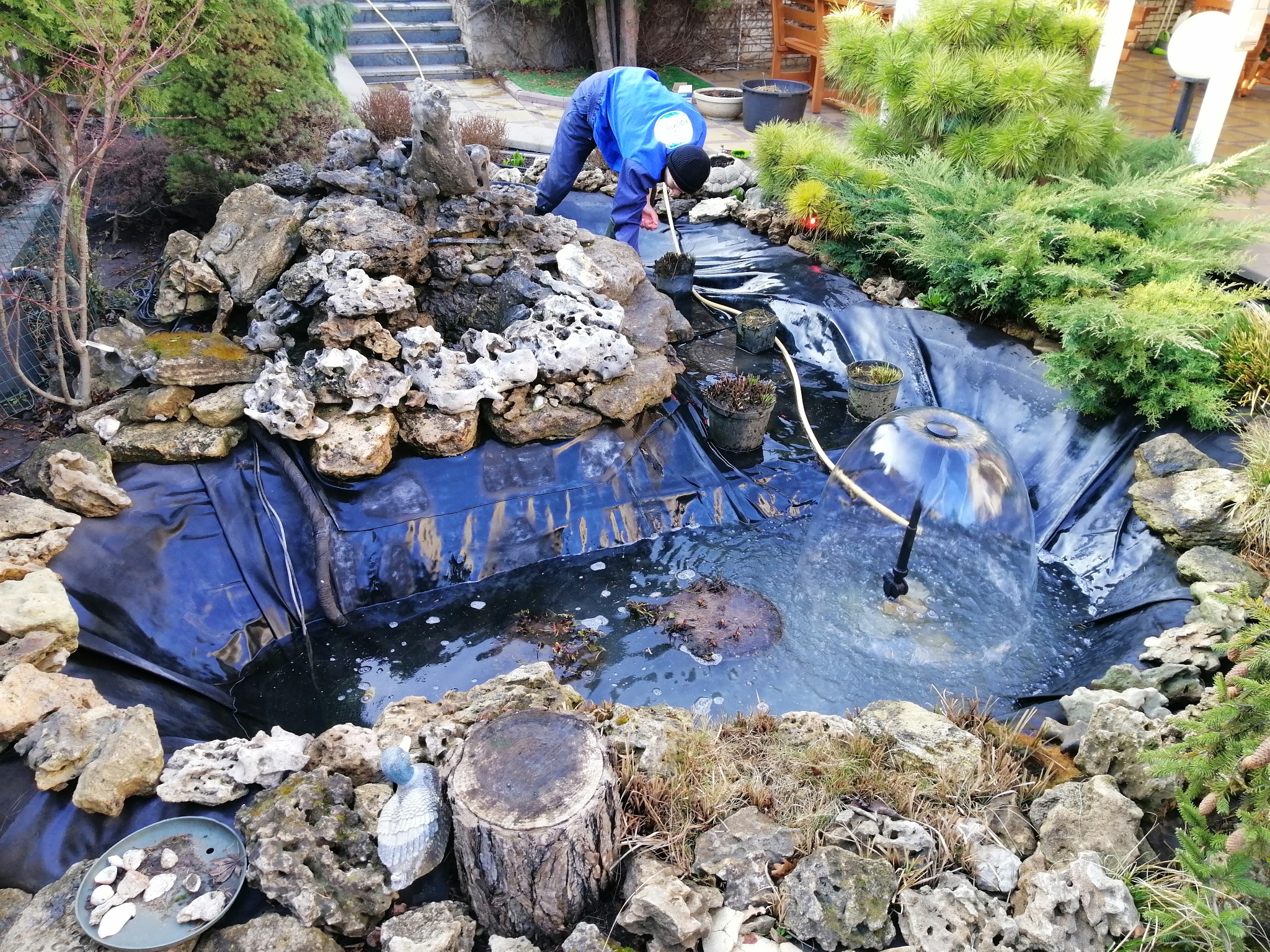 Prace wykończeniowe wokół oczka wodnego: układanie kamieni, sadzenie roślin. W tle schody i roślinność ogrodowa.