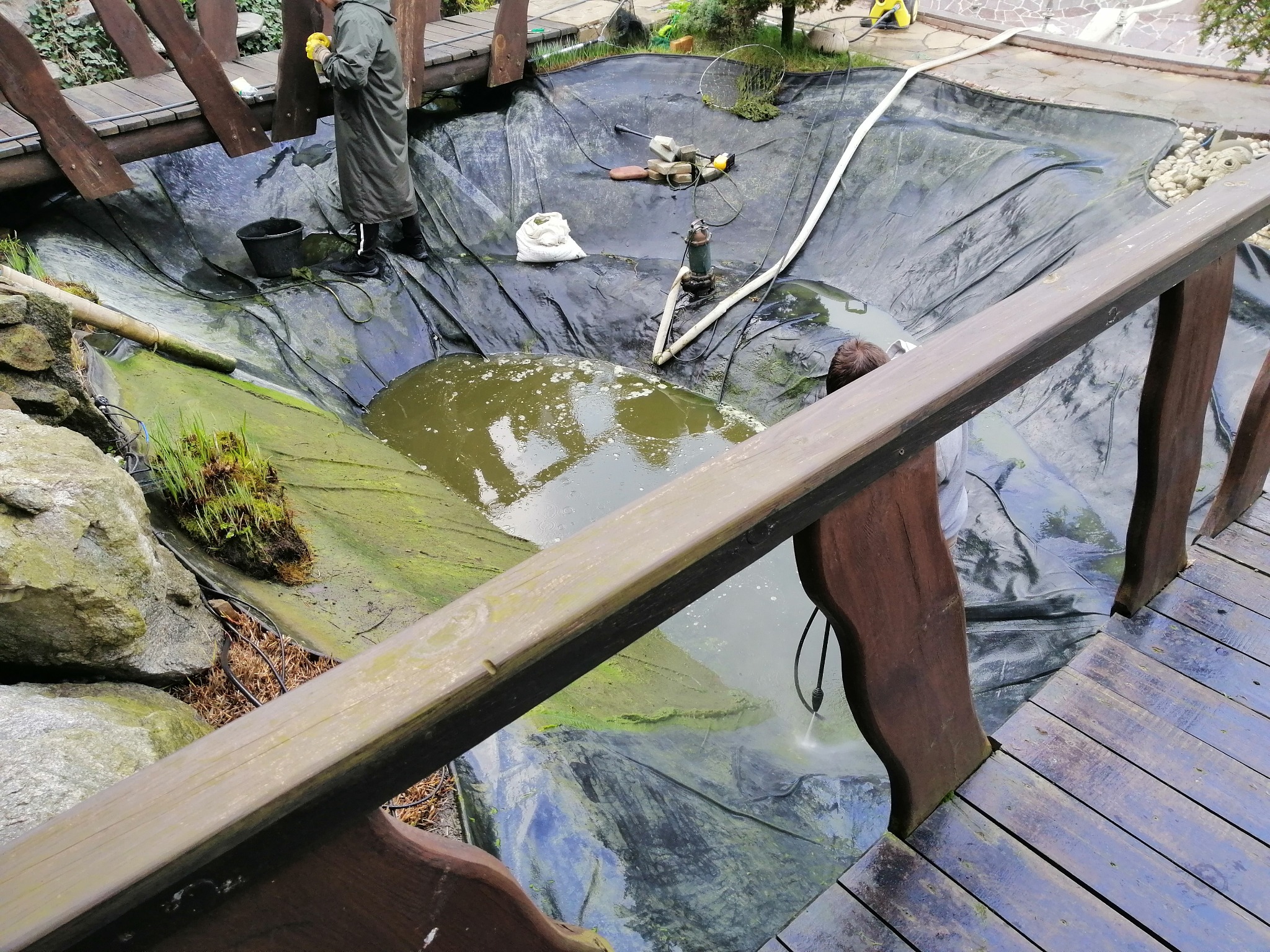Czyszczenie oczka wodnego z drewnianego mostku: osoba w płaszczu, pompa, narzędzia, woda z algami, czarna folia. Widok z góry, częściowe ujęcie.