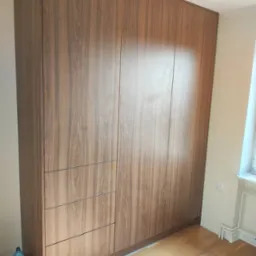 Wysoka, zabudowana szafa wnękowa z drewnopodobnymi frontami, podzielona na segment z trzema szufladami i część drzwiową. Butelka wody przy podstawie szafy.