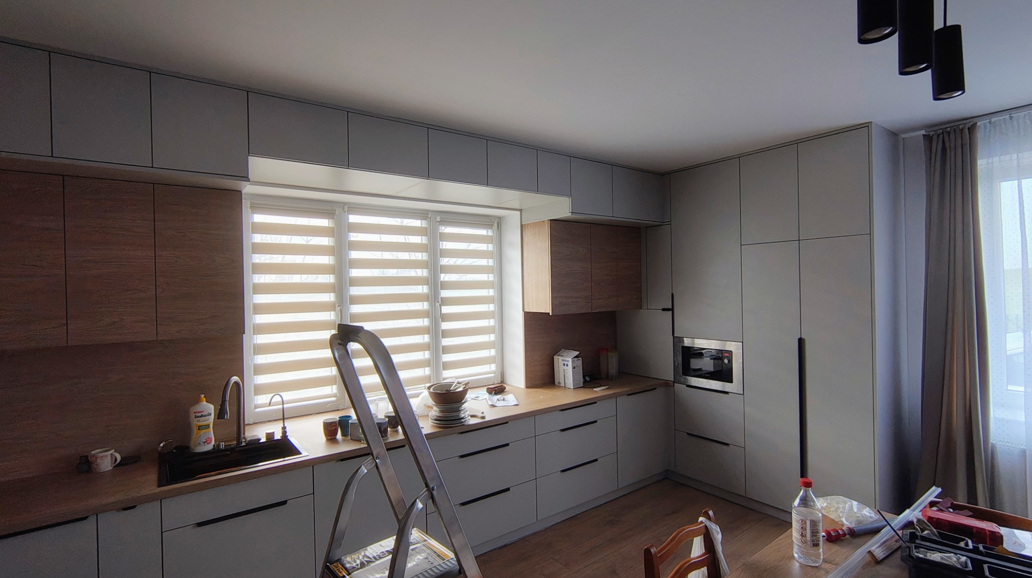 Jasnoszara kuchnia z drewnianymi akcentami, widok z drabiny, okno z roletami dzień-noc, częściowo widoczne narzędzia, podłoga drewniana, nowoczesne oświetlenie.