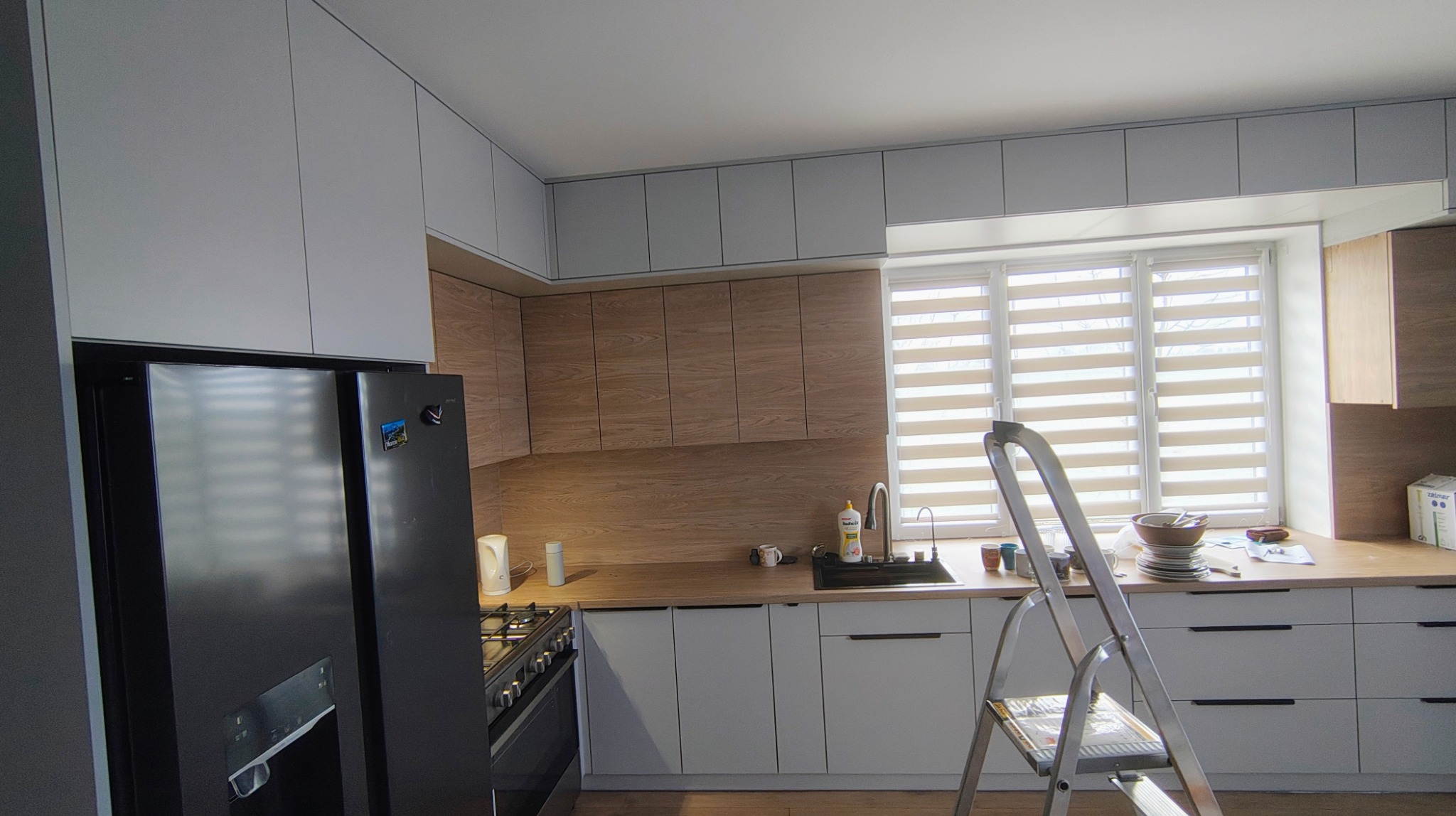 Jasna kuchnia z drewnianymi elementami, widziana z perspektywy podłogi. Na pierwszym planie drabina. Nowoczesne szafki wiszące i stojące.