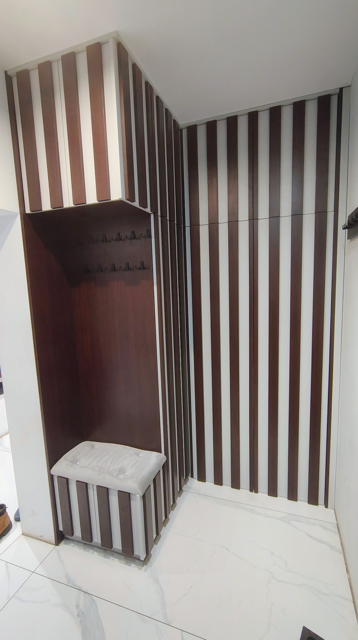 Nowoczesna zabudowa przedpokoju z siedziskiem i wieszakami, wykończona pionowymi panelami w odcieniach brązu i bieli, na tle marmurowej posadzki.