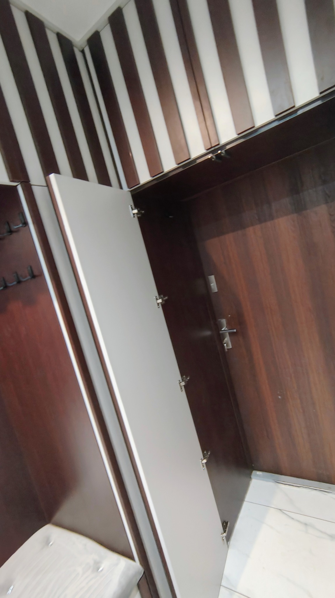 Otwarta szafa wnękowa z ciemnego drewna i białymi frontami, widoczne zawiasy i wnętrze. Na ścianie dekoracyjne pasy w kolorze drewna i bieli.