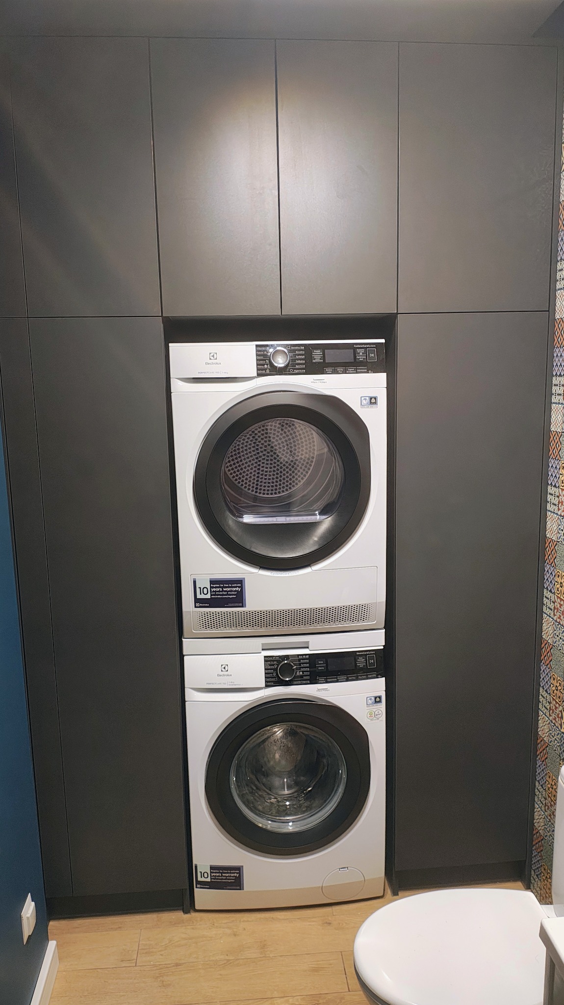 Szara zabudowa meblowa z pralką i suszarką Electrolux ustawionymi jedna na drugiej. Widoczna toaleta i fragment wzorzystej ściany.